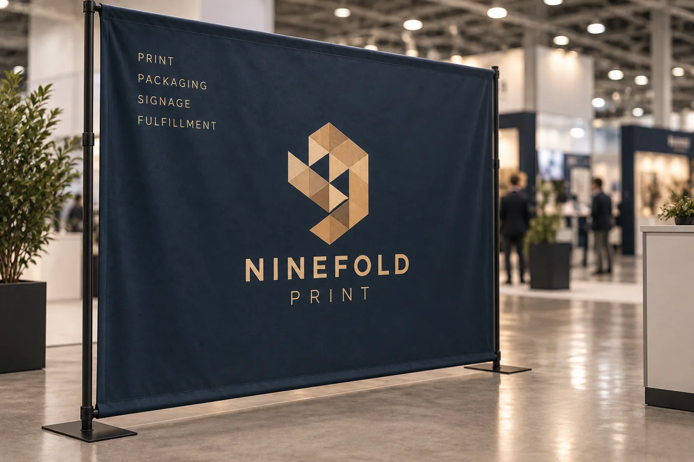 A tensioned fabric banner on a black stand in a trade show hall, deep navy with a large gold mark and a short service list
